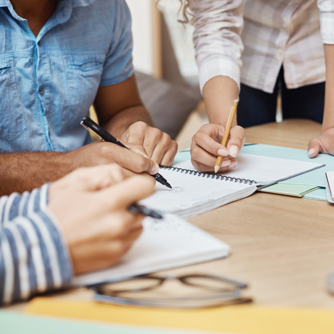 Manos de dos personas tomando notas con lápiz sobre una libreta en una mesa de trabajo.